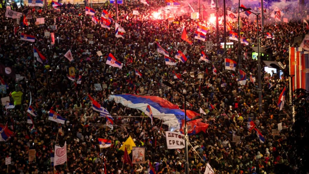 A vast crowd of demonstrators fills a Belgrade boulevard at night, waving Serbian flags and flares.