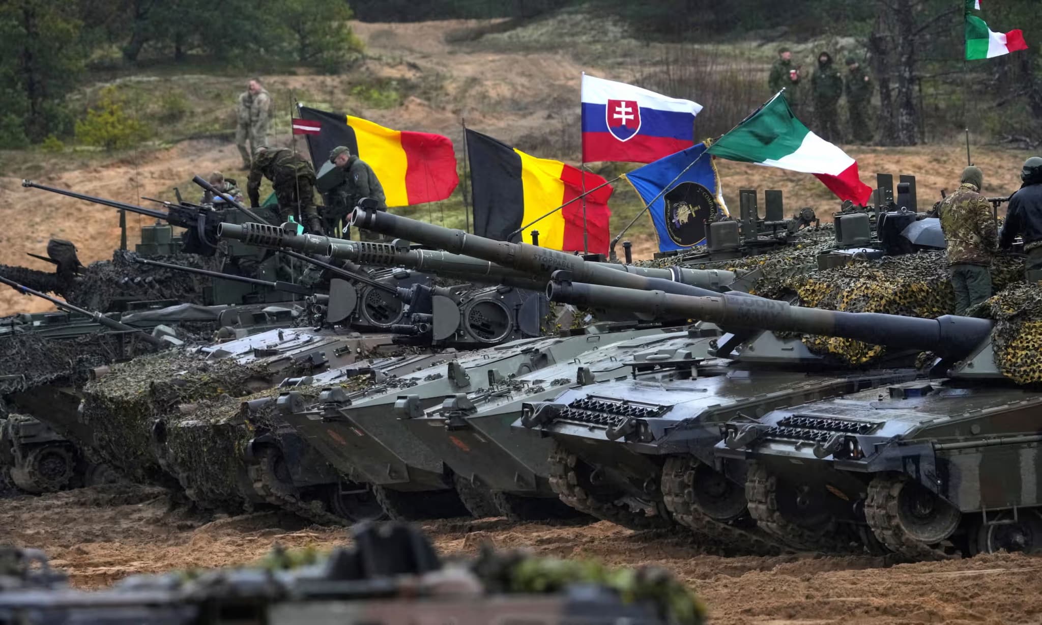 NATO armoured vehicles flying allied national flags during a live fire exercise in Latvia.